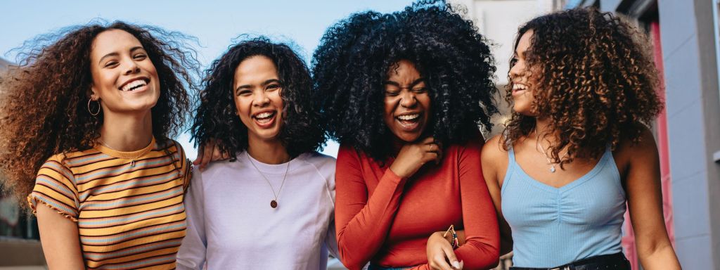 Four women with curly hair laughing and enjoying each other's company outdoors.